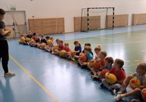 Prowadząca zaajęcia tłumaczy dzieciom w jaki sposób kozłować piłkę