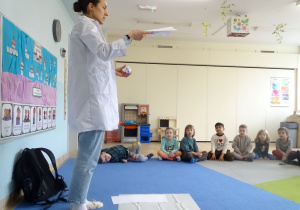 dzieci siedzą i patrzą na prowadzącą zajęcia która trzyma kartki