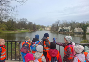 Dzieci stoją na mostku, trzymają maskotkę, w oddali widać Pałac na Wyspie