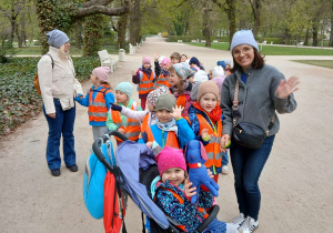 Dzieci z nauczycielem stoją na parkowej alejce i machają